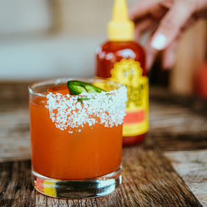 Little Birdie Cocktail in a glass, garnished with a jalapeño coin and a salted rim, with a bottle of Yellowbird Jalapeño Hot Sauce in the background