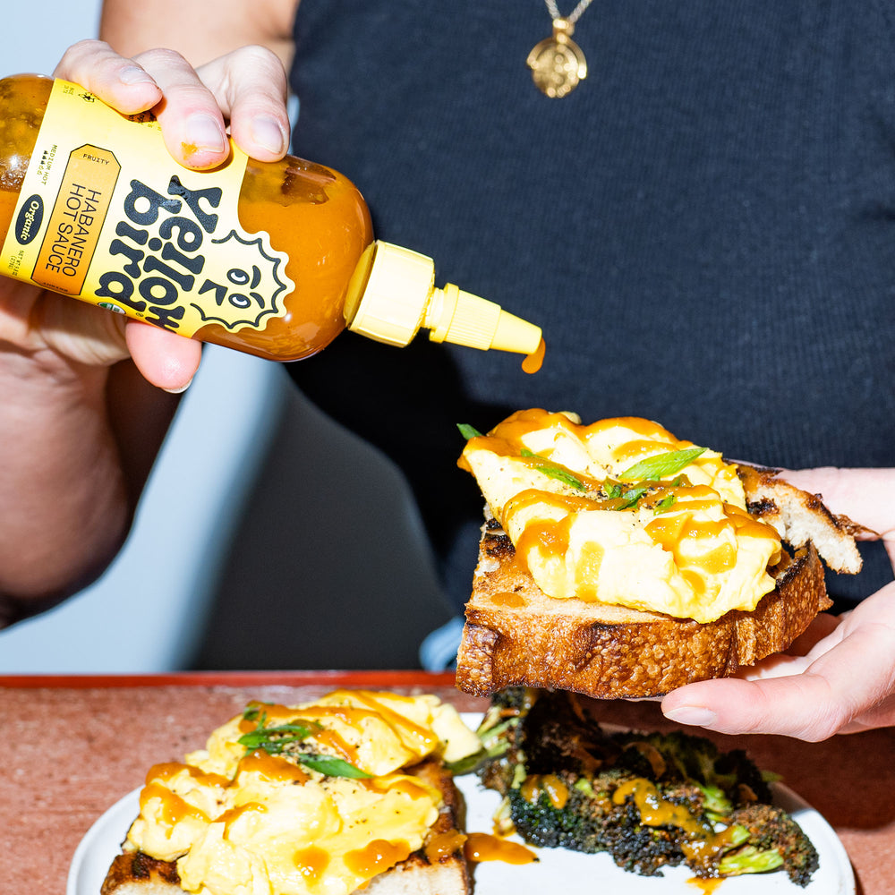Person pouring Yellowbird Organic Habanero Hot Sauce onto fluffy scrambled egg toast with with crispy broccoli.