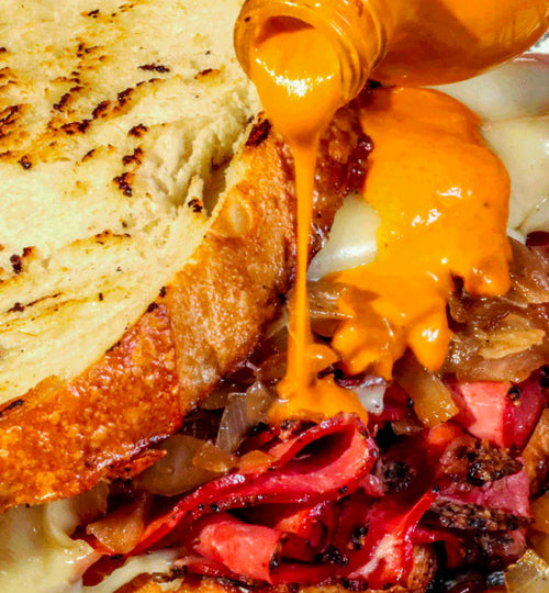 A picture of Yellowbird habanero hot sauce being drizzled on an open pastrami deli sandwich 