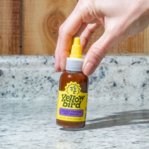 A picture of a Yellowbird Classic Ghost Pepper Hot Sauce in the 2.2  ounce size being held up by a persons fingers